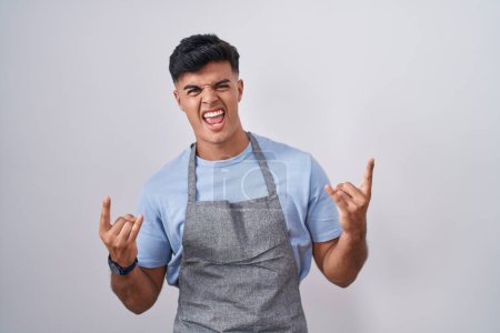 Photo for Hispanic young man wearing apron over white background shouting with crazy expression doing rock symbol with hands up. music star. heavy music concept. - Royalty Free Image