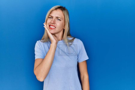 Photo for Beautiful blonde woman wearing casual t shirt over blue background touching mouth with hand with painful expression because of toothache or dental illness on teeth. dentist - Royalty Free Image