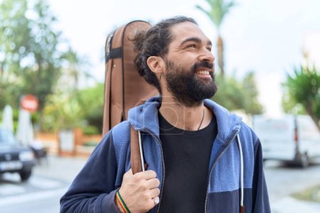 Foto de Joven músico hispano sonriendo confiado sosteniendo estuche de guitarra en la calle - Imagen libre de derechos
