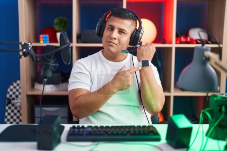 Téléchargez les photos : Jeune homme hispanique jouant à des jeux vidéo à la hâte pointant vers regarder le temps, l'impatience, regardant la caméra avec une expression détendue - en image libre de droit