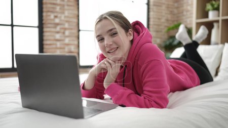 Téléchargez les photos : Jeune femme blonde utilisant un ordinateur portable couché sur le lit dans la chambre - en image libre de droit