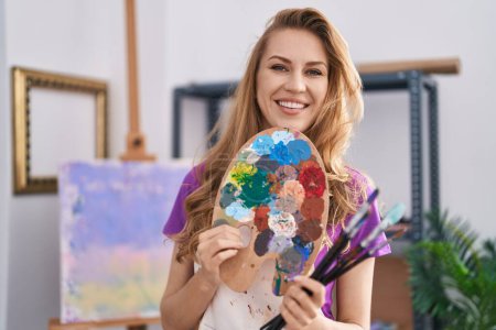 Photo for Young blonde woman artist smiling confident holding paintbrushes and palette at art studio - Royalty Free Image