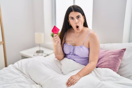 Téléchargez les photos : Jeune femme brune portant de la lingerie et tenant un préservatif sur le lit effrayé et étonné avec la bouche ouverte pour la surprise, visage incrédule - en image libre de droit