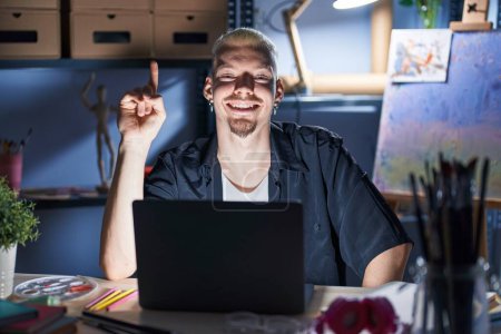 Foto de Joven hombre caucásico usando el ordenador portátil por la noche en el estudio de arte mostrando y señalando con el dedo número uno mientras sonríe confiado y feliz. - Imagen libre de derechos