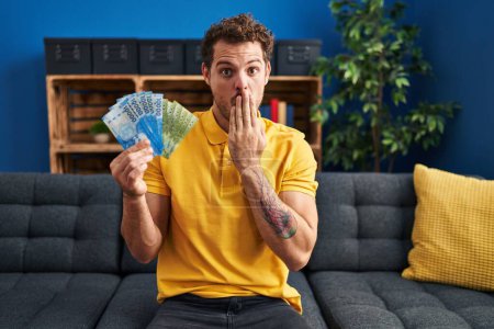 Photo for Young hispanic man holding chilean pesos covering mouth with hand, shocked and afraid for mistake. surprised expression - Royalty Free Image