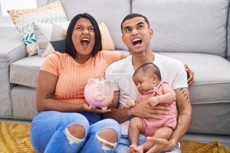 Photo for Young hispanic couple with baby holding piggy bank angry and mad screaming frustrated and furious, shouting with anger looking up. - Royalty Free Image