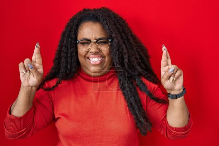 Photo for Plus size hispanic woman standing over red background gesturing finger crossed smiling with hope and eyes closed. luck and superstitious concept. - Royalty Free Image