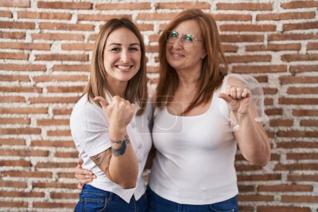 Téléchargez les photos : Mère et fille hispaniques portant un t-shirt blanc décontracté pointant vers le dos derrière avec la main et les pouces levés, souriant confiant - en image libre de droit