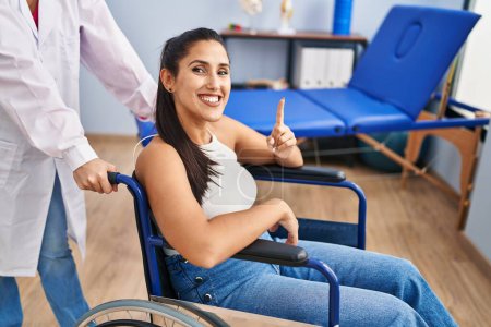 Photo for Young hispanic woman sitting on wheelchair at physiotherapy clinic surprised with an idea or question pointing finger with happy face, number one - Royalty Free Image
