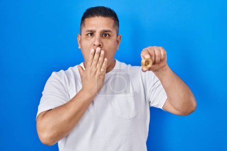 Téléchargez les photos : Jeune homme hispanique tenant monnaie virtuelle Bitcoin couvrant la bouche avec la main, choqué et peur de l'erreur. expression surprise - en image libre de droit