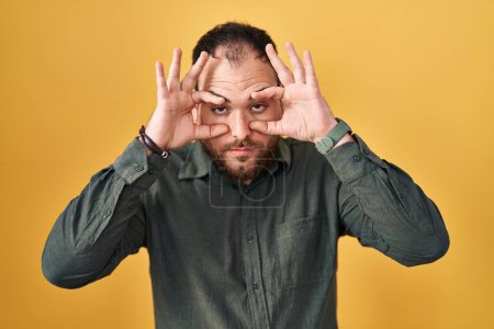 Téléchargez les photos : Homme hispanique de taille plus avec barbe debout sur fond jaune essayant d'ouvrir les yeux avec les doigts, somnolent et fatigué pour la fatigue matinale - en image libre de droit