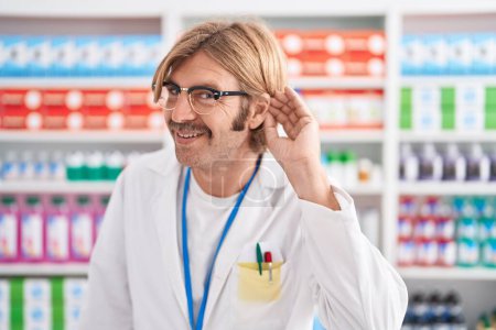 Foto de Hombre caucásico con bigote trabajando en farmacia sonriendo con la mano sobre el oído escuchando rumores o chismes. concepto de sordera. - Imagen libre de derechos