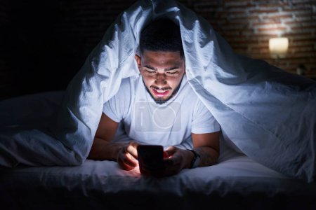 Téléchargez les photos : Young latin man using smartphone lying on bed at bedroom - en image libre de droit