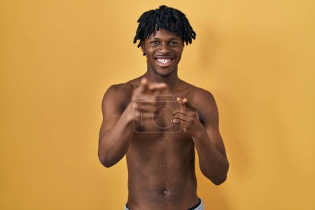 Foto de Joven hombre africano con rastas de pie sin camisa señalando los dedos a la cámara con la cara feliz y divertida. buena energía y vibraciones. - Imagen libre de derechos