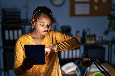 Téléchargez les photos : Femme afro-américaine avec des tresses travaillant au bureau la nuit avec tablette regardant l'heure de la montre inquiète, peur de se retrouver en retard - en image libre de droit