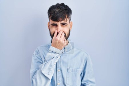 Téléchargez les photos : Jeune homme hispanique avec la barbe debout sur fond bleu sentant quelque chose de puant et dégoûtant, odeur intolérable, retenant souffle avec les doigts sur le nez. mauvaise odeur - en image libre de droit