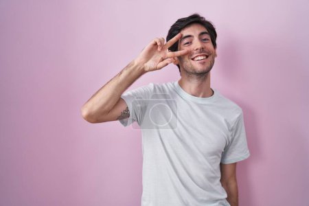 Téléchargez les photos : Jeune homme hispanique debout sur fond rose faisant symbole de paix avec les doigts sur le visage, souriant joyeux montrant la victoire - en image libre de droit