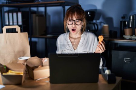 Photo for Young beautiful woman working using computer laptop and eating delivery food in shock face, looking skeptical and sarcastic, surprised with open mouth - Royalty Free Image