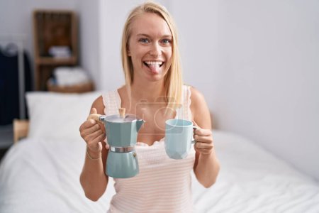 Foto de Joven mujer caucásica bebiendo café en la cama sacando la lengua feliz con expresión divertida. - Imagen libre de derechos
