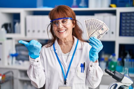 Téléchargez les photos : Moyen Age femme hispanique travaillant au laboratoire scientifique tenant des dollars pointant pouce vers le côté souriant heureux avec la bouche ouverte - en image libre de droit