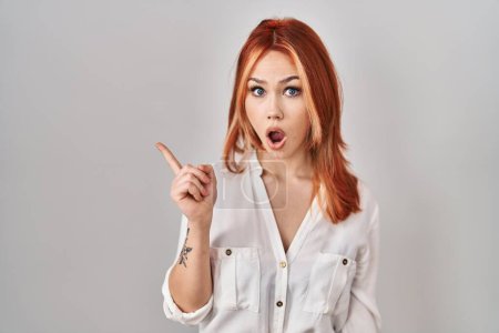 Photo for Young caucasian woman standing over isolated background surprised pointing with finger to the side, open mouth amazed expression. - Royalty Free Image
