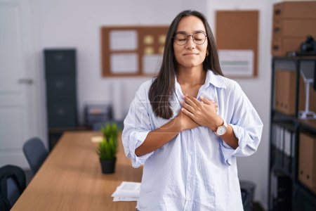 Foto de Mujer hispana joven en la oficina sonriendo con las manos en el pecho con los ojos cerrados y gesto agradecido en la cara. concepto de salud. - Imagen libre de derechos