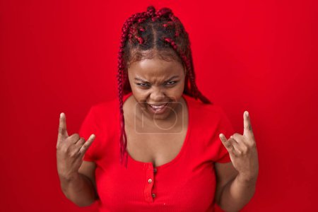 Téléchargez les photos : Femme afro-américaine avec des cheveux tressés debout sur fond rouge criant avec une expression folle faire symbole rock avec les mains en l'air. star de la musique. concept lourd. - en image libre de droit