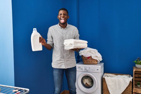 Foto de Hombre afroamericano sosteniendo toallas limpias sacando la lengua feliz con expresión divertida. - Imagen libre de derechos