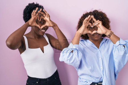 Téléchargez les photos : Deux femmes africaines debout sur fond rose faisant forme de coeur avec la main et les doigts souriant en regardant à travers signe - en image libre de droit