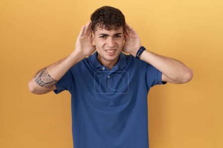 Photo for Young hispanic man standing over yellow background trying to hear both hands on ear gesture, curious for gossip. hearing problem, deaf - Royalty Free Image