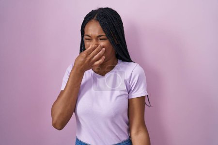 Foto de Mujer afroamericana con trenzas de pie sobre fondo rosa oliendo algo apestoso y asqueroso, olor intolerable, conteniendo la respiración con los dedos en la nariz. mal olor - Imagen libre de derechos