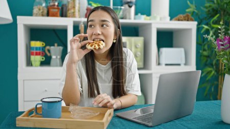 Téléchargez les photos : Jeune belle femme hispanique prenant le petit déjeuner à l'aide d'un ordinateur portable à la salle à manger - en image libre de droit