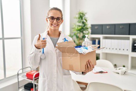 Foto de Mujer doctora joven sosteniendo caja con artículos médicos sonriendo feliz y positivo, pulgar hacia arriba haciendo excelente y signo de aprobación - Imagen libre de derechos