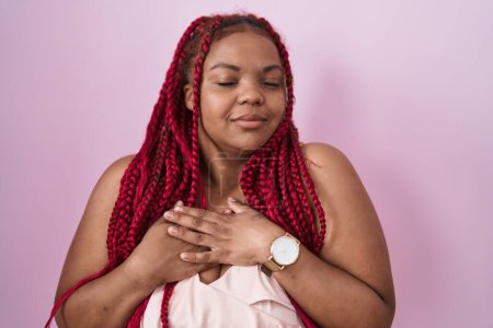 Téléchargez les photos : Femme afro-américaine avec les cheveux tressés debout sur fond rose souriant avec les mains sur la poitrine avec les yeux fermés et geste reconnaissant sur le visage. concept de santé. - en image libre de droit