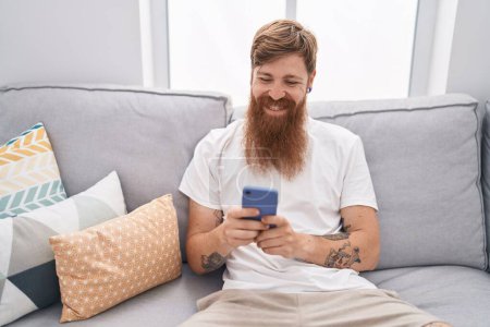 Photo for Young redhead man using smartphone sitting on sofa at home - Royalty Free Image