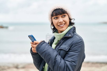 Foto de Joven hermosa mujer hispana sonriendo confiada usando teléfono inteligente en la playa - Imagen libre de derechos