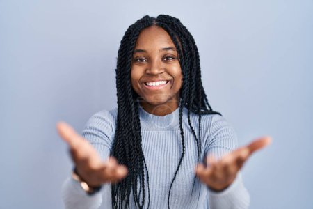 Foto de African american woman standing over blue background smiling cheerful offering hands giving assistance and acceptance. - Imagen libre de derechos