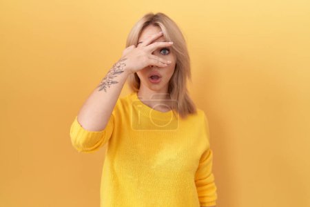 Foto de Joven mujer caucásica con suéter amarillo asomándose en shock cubriendo la cara y los ojos con la mano, mirando a través de los dedos con expresión avergonzada. - Imagen libre de derechos