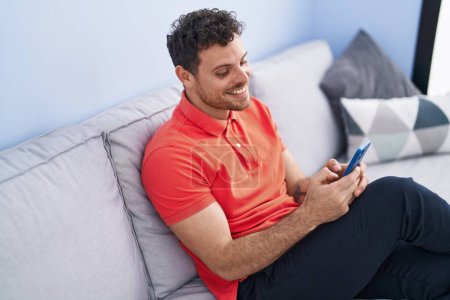 Téléchargez les photos : Jeune homme hispanique utilisant smartphone assis sur le canapé à la maison - en image libre de droit