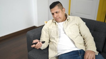 Photo for Young hispanic man watching tv sitting on sofa at home - Royalty Free Image