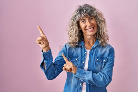 Photo for Middle age woman standing over pink background smiling and looking at the camera pointing with two hands and fingers to the side. - Royalty Free Image