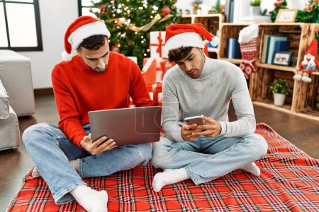 Foto de Dos hombres hispanos en pareja usando laptop y smartphone sentados junto al árbol de navidad en casa - Imagen libre de derechos