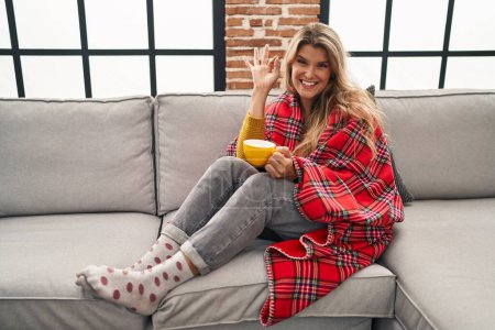 Foto de Mujer joven sentada en el sofá bebiendo un café en casa sonriendo positiva haciendo signo de ok con la mano y los dedos. expresión exitosa. - Imagen libre de derechos