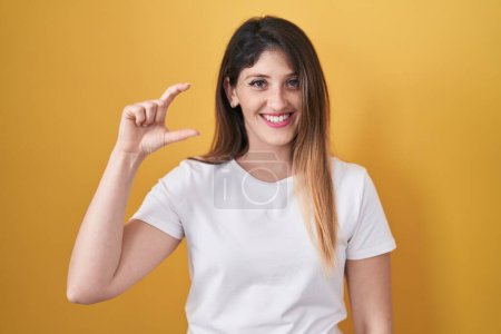 Téléchargez les photos : Jeune femme brune debout sur fond jaune geste souriant et confiant avec la main faisant signe de petite taille avec les doigts regardant et la caméra. concept de mesure. - en image libre de droit