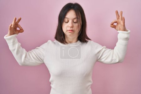 Téléchargez les photos : Femme avec le syndrome du duvet debout sur fond rose détendue et souriante avec les yeux fermés faisant geste de méditation avec les doigts. concept de yoga. - en image libre de droit