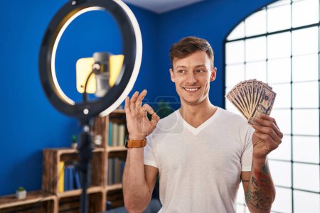 Photo for Caucasian man recording vlog tutorial with smartphone at home holding money doing ok sign with fingers, smiling friendly gesturing excellent symbol - Royalty Free Image