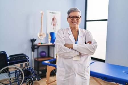 Téléchargez les photos : Femme d'âge moyen physiothérapeute debout avec les bras croisés geste à la clinique de réadaptation - en image libre de droit