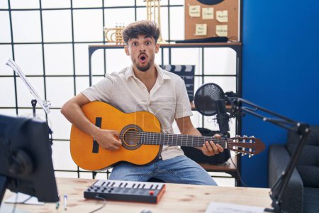 Photo for Arab man with beard playing classic guitar at music studio scared and amazed with open mouth for surprise, disbelief face - Royalty Free Image