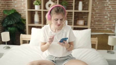 Téléchargez les photos : Young blonde woman listening to music sitting on bed at bedroom - en image libre de droit