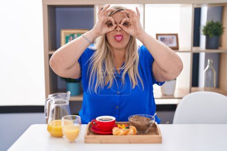 Foto de Mujer caucásica de talla grande desayunando en casa haciendo un buen gesto como binoculares sacando la lengua, ojos mirando a través de los dedos. expresión loca. - Imagen libre de derechos